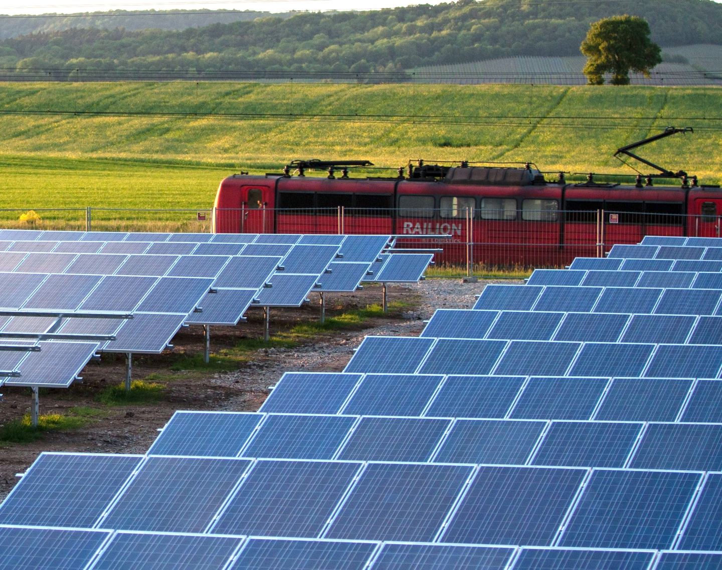 Solar cells in the foreground with a red train passing behind them, and grassy field and trees in the background.